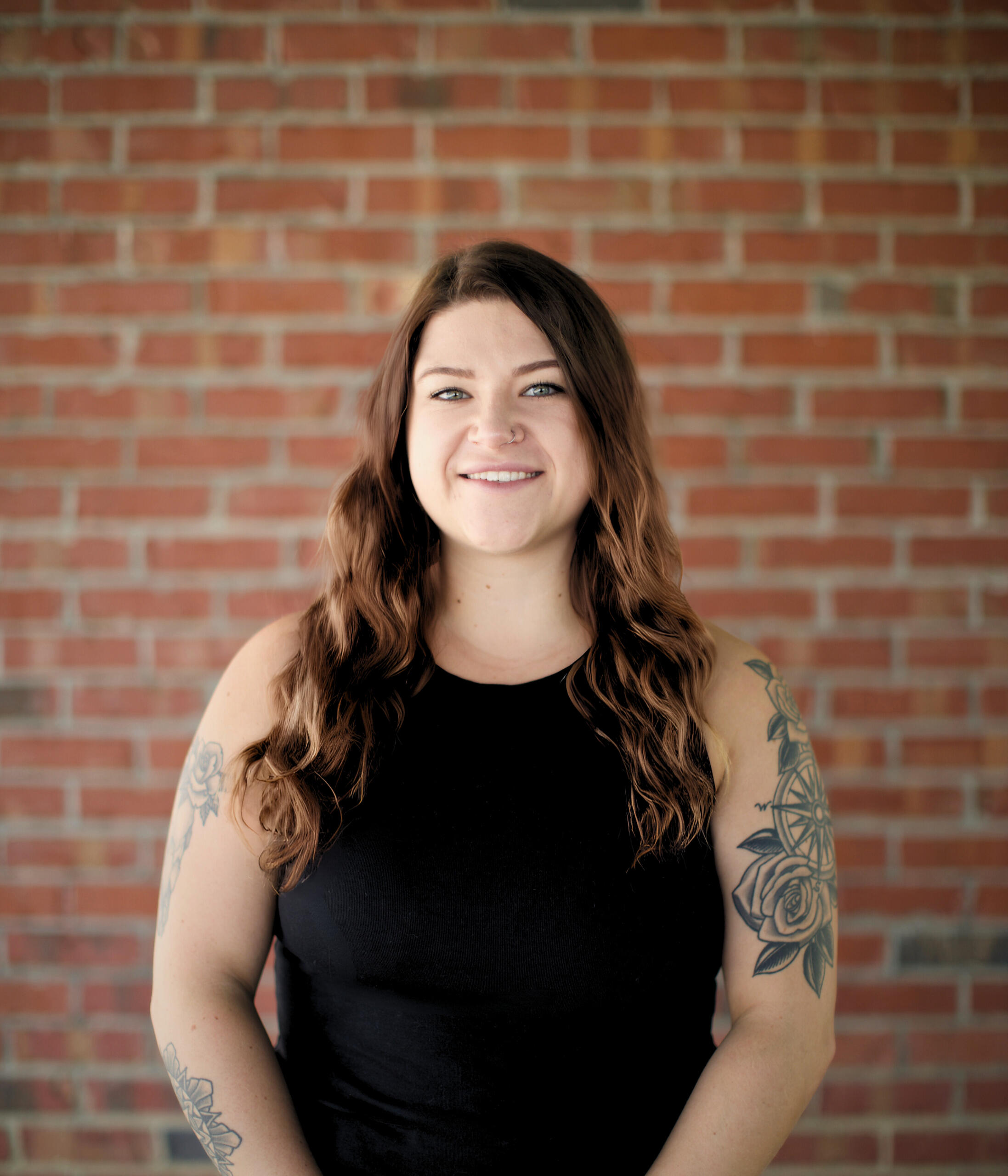 Kat Stoner | REALTOR® Kat Stoner, a smiling real estate agent with long wavy hair and visible tattoos, standing in front of a brick wall.