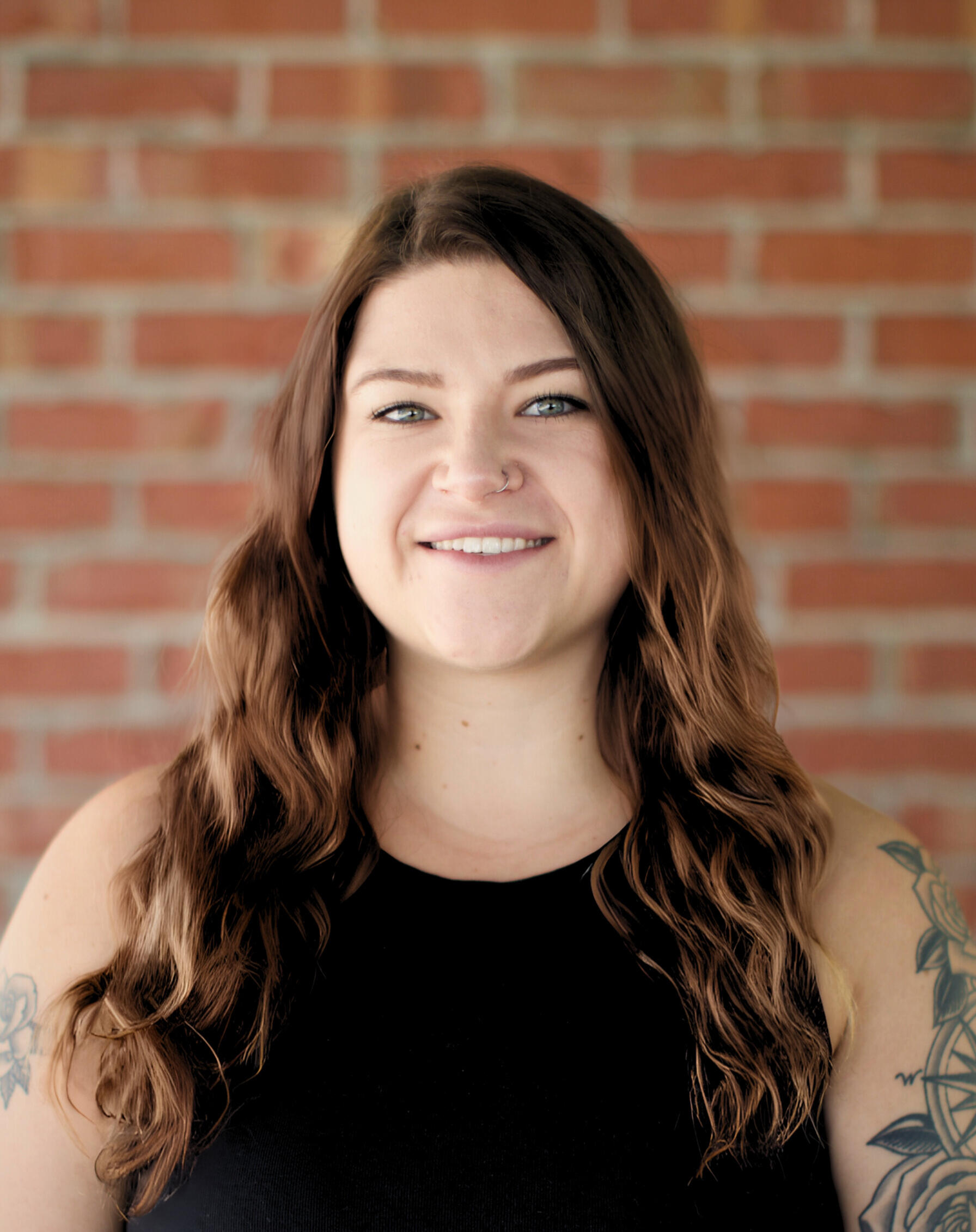 Kat Stoner | REALTOR® Kat Stoner, a smiling real estate agent with long wavy hair and visible tattoos, standing in front of a brick wall.